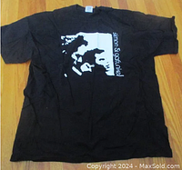 Full view of the black T-shirt laid flat on a wooden floor showing the graphic design with Simon & Garfunkel faces.
