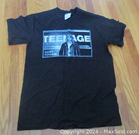 Black t-shirt laid flat on wood floor with a large rectangular black and white graphic of Kurt Cobain and text 'TEENAGE' on front.