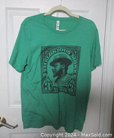 Full view of the green T-shirt hanging on a hanger, showing the black graphic print of Thelonious Monk on the front.