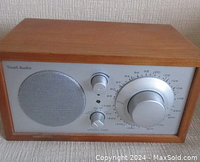 Front view of Tivoli Audio Model One radio showing walnut wood frame, silver front panel with speaker grille, knobs for power/source and volume, and large tuning dial.