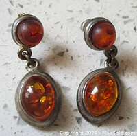 Pair of vintage cognac Baltic amber earrings on a light speckled surface. Each earring has a round amber stone on top and a larger oval amber stone below. Metal settings slightly tarnished.