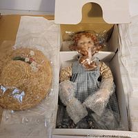 Photo of Anne Shirley doll in original box with protective plastic on arms and legs, alongside the straw hat in plastic wrapping.