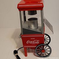 Side view of Coca-Cola hot air popcorn maker showing red metal body, black wheels, glass panels, aluminum popping chamber, and power cord.