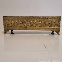 Front view of the rectangular copper flower holder showing detailed raised leaf and vine design and four ornate feet.