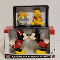 Front view of two boxed ceramic salt and pepper shaker sets: one featuring Winnie The Pooh with a honey pot, and another featuring Mickey Mouse and Minnie Mouse kissing.