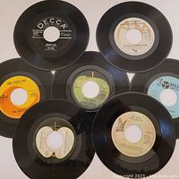 Top view showing seven 45 RPM vinyl records spread out, featuring labels from The Beatles, The Who, John Lennon, Peter Frampton, and The Cars.