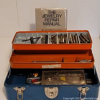 Blue metal toolbox with open lid showing orange tray filled with metal taps and dies, and jewelry repair manual standing at the back