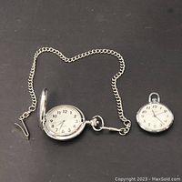 Full view of two silver-colored pocket watches, one with chain, both showing white dials with black numerals and hands on dark background