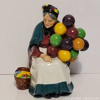 Front view of the Royal Doulton The Old Balloon Seller figurine showing the lady holding colorful balloons and basket seated at her side.