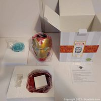 Photo showing the Iron Man helmet warmer wrapped in plastic, original foam packaging, box with Marvel branding and instructional paper.
