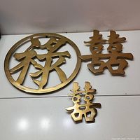 Three brass wall decor pieces set on a white surface, including one circular character and two 'Double Happiness' characters.
