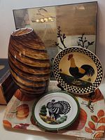 Photo showing a brown swirl patterned ceramic vase, a rooster porcelain plate with checkered rim, a turkey porcelain plate on a plastic autumn harvest tray, and an autumn-themed picture behind them.