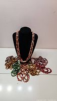 Photo showing eight bead necklaces draped on and around a black display stand. The beads have irregular shapes and reflective surfaces in various colors including greens, browns, reds, and golds.