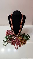 Seven bead necklaces arranged with one orange necklace displayed on a black bust and the other six laid beside it in different colors.