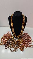 Photo showing one long square amber-colored beaded necklace displayed on a black mannequin bust and eight additional necklaces in various earthy tones laid around.