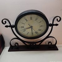 Front angled view of the decorative clock on a flat surface showing the ornate metal bracket stand and Roman numeral dial.