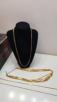 Gold-colored chain displayed on a black necklace stand and several chains laid flat on a white surface.