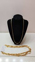 Photo of a single gold-tone necklace on display bust with smooth chain, and multiple necklaces stacked below