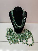 Multiple green and clear bead necklaces displayed on a black stand with four necklaces spread on the table below.