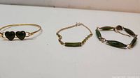 Photo showing three bracelets: a gold-tone bracelet with two dark green heart-shaped jade stones, a delicate chain bracelet with small green jade beads and a central rectangular jade element, and a bracelet with elongated dark green jade links connected by metal.