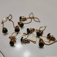 Close-up of several jade and gold-tone jacket pins with oval attachments connected by fine chains, showing the green jade stones, gold-tone metal setting, and chain details.