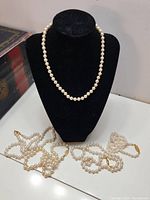 One medium length pearl necklace displayed on a black velvet bust with four additional pearl necklaces laid out on a white surface nearby.
