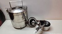 Photo of stainless steel 2 compartment double wall tiffin box with handle, four nested measuring cups, and manual citrus juicer arranged on table.