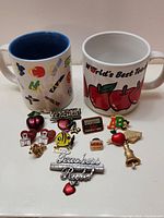 Two ceramic teacher-themed coffee mugs with 11 assorted teacher pins displayed in front.
