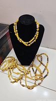 Five yellow bead necklaces displayed on a stand and laid out on a table, showing mixed beads and cylindrical elements.