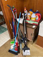Photo showing a collection of floor cleaning tools leaning against a wall and a small metal cabinet with various cleaning liquids and products on top.