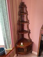 Full view of wooden corner display shelf showing five shelves and bottom cabinet in corner near window and curtain.