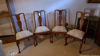 Four vintage wood dining chairs with brocade cushioned seats arranged side by side on carpeted floor.