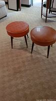 Two vintage round orange vinyl foot stools on carpeted floor, both with wooden legs and brass feet caps. Visible wear, one leg loose.