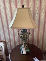 Single bulb table lamp with ornate base and fabric shade shown from front angle on round wooden table against patterned wallpaper.