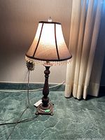 Full view of the table lamp lit, showcasing the beige fabric shade with bead trim and carved decorative wooden base.