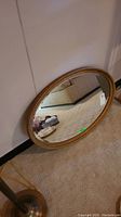 Oval mirror leaning against wall on carpeted floor, showing full mirror and frame.