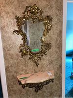 Ornate gold gilt framed wall mirror hung above matching pink marble topped shelf with decorative metal bracket.