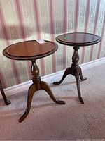 Pair of matching three-legged wooden tables with round tops and turned pedestals on carpeted floor against wallpapered wall.