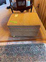 Front-left side view of the beige upholstered ottoman showing textured fabric and castors.