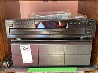 Sony CDP-CE375 CD player sitting atop a black rectangular storage case inside a wooden shelving unit. The player has various buttons and display on front.