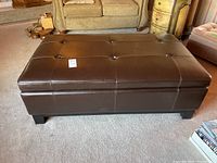Front view showing brown faux leather tufted ottoman with black block legs on carpeted floor.