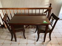 View of vintage wooden dining table with custom glass top and two harp back chairs against a bannister wall with patterned wallpaper and tiled floor.