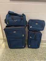 Four blue luggage pieces including two rolling suitcases, a tote bag, and a shoulder carry bag, arranged on a floor near a wall.