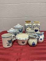 Nine-piece blue and white ceramic kitchenware set with heart and floral designs arranged on a red cloth.