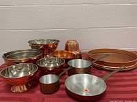 12 pieces of copper kitchenware arranged on maroon cloth, includes pans, pots, nesting bowls, colanders, spring form pan, and jello mould.