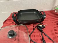 Electric fry pan with glass lid and temperature control cord placed on a red cloth.