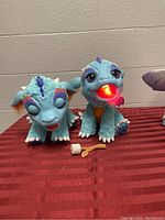 Two blue interactive Furreal Friends Torch My Blazin Dragon toys on red striped surface with marshmallow roaster accessory in front.