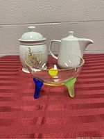 Photo showing ceramic canister, white ceramic teapot, and colorful glass bowl on a red striped surface