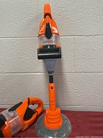Front view of the Home Depot kids toy weed eater standing upright on a red surface against a white wall. The toy is orange with gray and clear plastic parts and has a circular base.