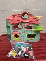 Playset with red roof and turquoise colored walls, showing front of hotel house with floral decorations and yellow door with 'Adventure' label. Bag of accessories in front on red tablecloth.